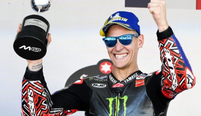 Motocross rider wearing a blue cap and sunglasses celebrates on stage, holding a trophy aloft and smiling broadly.