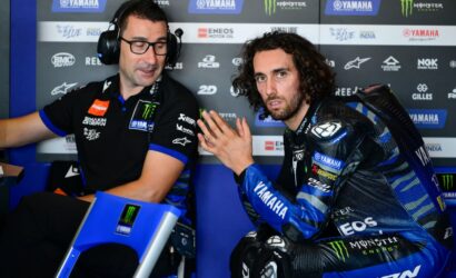 Two men in a Yamaha pit area: a rider in blue racing gear sits, talking with a crew member wearing a headset.