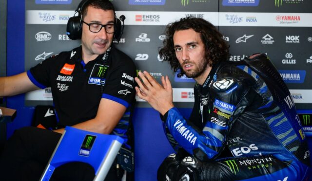 Two men in a Yamaha pit area: a rider in blue racing gear sits, talking with a crew member wearing a headset.