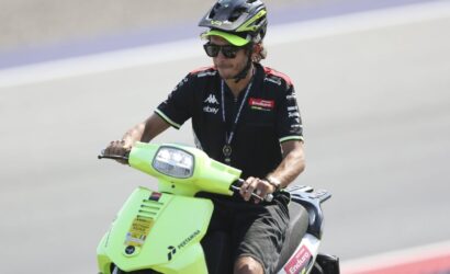 Man wearing a helmet and sunglasses rides a bright lime-green scooter on a race track with sponsor logos on his shirt and the bike front plate visible.