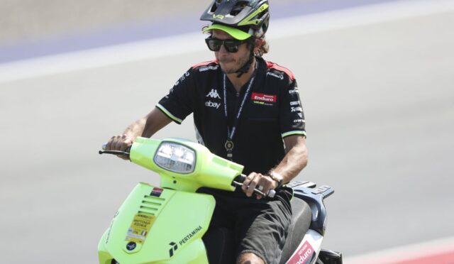 Man wearing a helmet and sunglasses rides a bright lime-green scooter on a race track with sponsor logos on his shirt and the bike front plate visible.