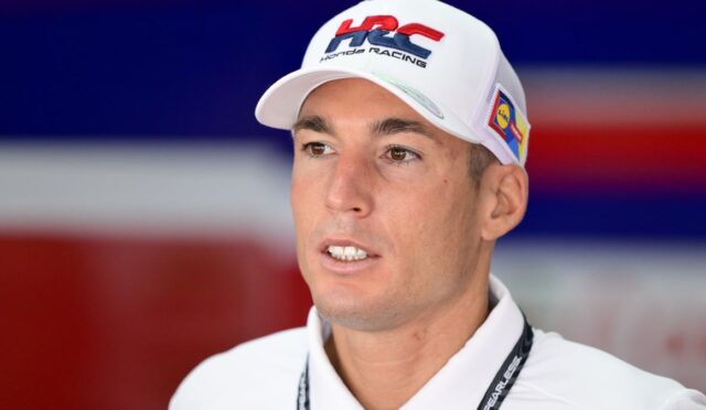 Portrait of a man wearing a white cap with HRC Honda Racing logo, speaking at a press event with a red-blue background.