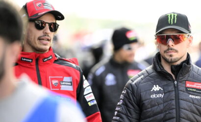 Two MotoGP riders in team gear stand in a paddock, one in red Ducati Lenovo suit and the other in a black jacket with sponsor patches.