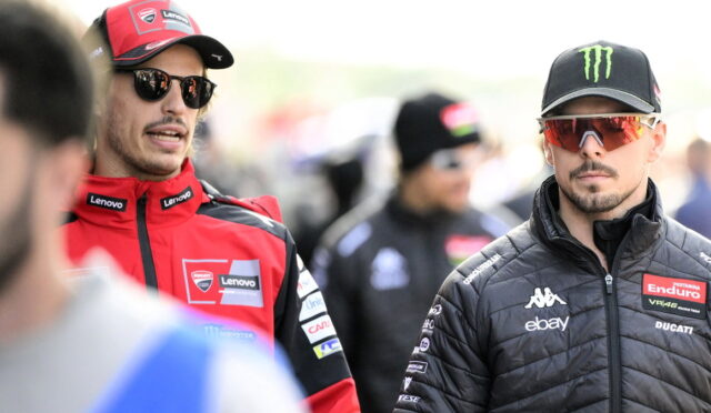 Two MotoGP riders in team gear stand in a paddock, one in red Ducati Lenovo suit and the other in a black jacket with sponsor patches.