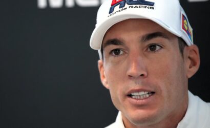 Close-up of a man in a white cap with racing branding, speaking at a press event, facing slightly right.