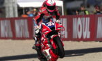 Moto racer in red-and-white gear performs a wheelie on a red Honda bike, number 51, on a race track with barriers behind him.