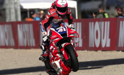 Moto racer in red-and-white gear performs a wheelie on a red Honda bike, number 51, on a race track with barriers behind him.