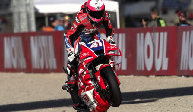 Moto racer in red-and-white gear performs a wheelie on a red Honda bike, number 51, on a race track with barriers behind him.