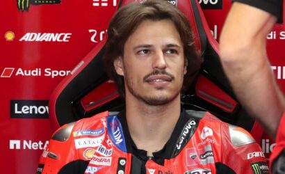 Male motorcycle racer in a red racing suit sits in a red and black sponsor-filled chair, looking off to the side in a garage/pit area.