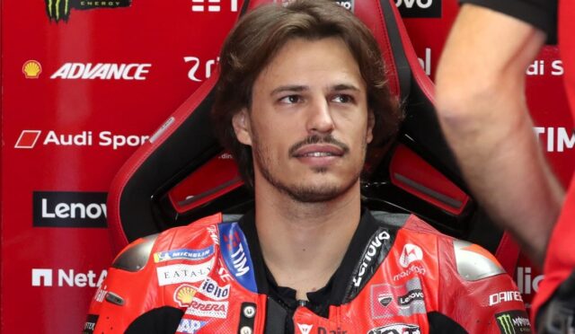 Male motorcycle racer in a red racing suit sits in a red and black sponsor-filled chair, looking off to the side in a garage/pit area.