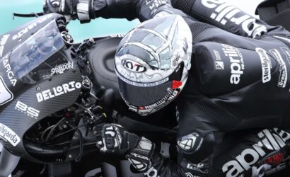 Motorcycle racer in black racing suit leans into a high-speed turn on a carbon-fiber race bike with sponsor logos visible on gear and bike.