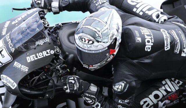 Motorcycle racer in black racing suit leans into a high-speed turn on a carbon-fiber race bike with sponsor logos visible on gear and bike.