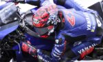 Professional motorcycle racer in a blue Yamaha suit crouches over a blue Yamaha race bike, inspecting the handlebars.
