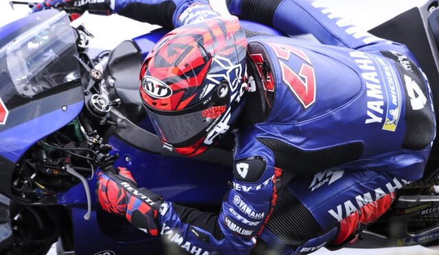 Professional motorcycle racer in a blue Yamaha suit crouches over a blue Yamaha race bike, inspecting the handlebars.