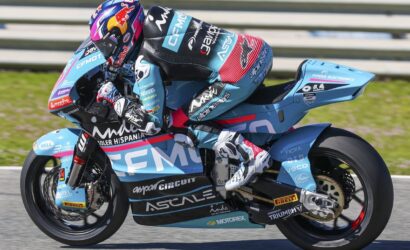 Blue racing motorcycle with a rider in a matching livery leaning into a turn on a race track, sponsor logos visible on the bike and suit.