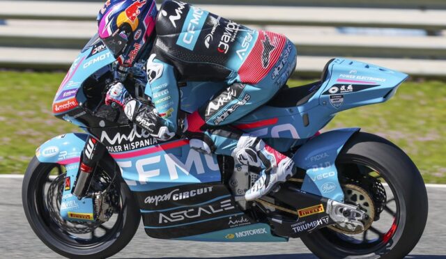 Blue racing motorcycle with a rider in a matching livery leaning into a turn on a race track, sponsor logos visible on the bike and suit.