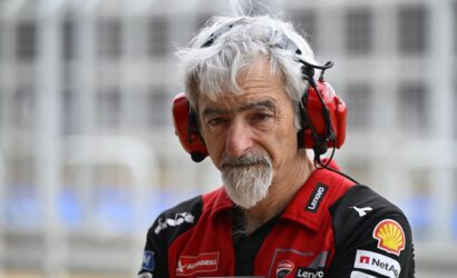 Professional race engineer wearing a red headset and red-black team shirt with sponsor logos, looking thoughtfully at the camera in the pit area.