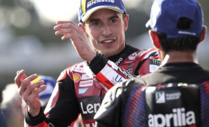 Racing rider in red and black gear with a Michelin cap smiles and gestures toward a teammate in a blue cap.