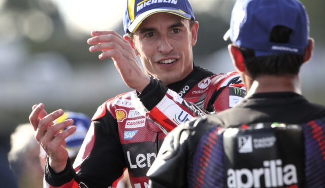 Racing rider in red and black gear with a Michelin cap smiles and gestures toward a teammate in a blue cap.