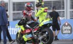 Two motorcycle racers in neon yellow and red racing suits talk beside a bright yellow bike on a track, with crew and a photographer nearby.