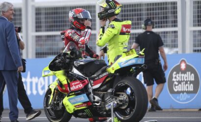 Two motorcycle racers in neon yellow and red racing suits talk beside a bright yellow bike on a track, with crew and a photographer nearby.