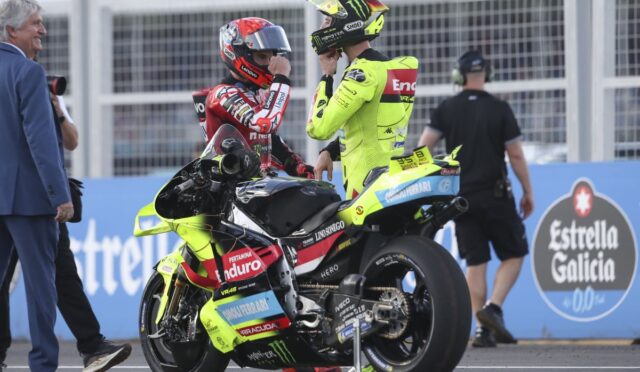 Two motorcycle racers in neon yellow and red racing suits talk beside a bright yellow bike on a track, with crew and a photographer nearby.