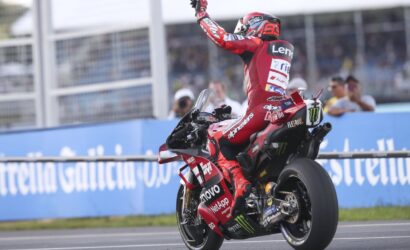 MotoGP rider in red racing suit performs a wheelie/victory pose on a red Ducati-like bike, sprinting on track with sponsor logos.