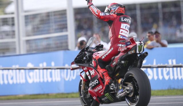 MotoGP rider in red racing suit performs a wheelie/victory pose on a red Ducati-like bike, sprinting on track with sponsor logos.