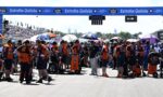 Pit crews in orange uniforms organize at the starting grid under an Estrella Galicia banner, with spectators behind them and race equipment scattered across the track.