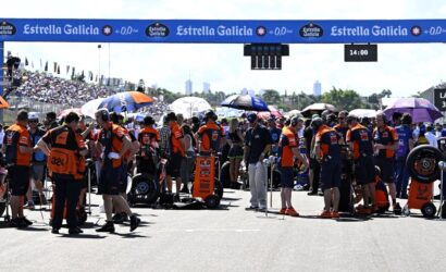 Pit crews in orange uniforms organize at the starting grid under an Estrella Galicia banner, with spectators behind them and race equipment scattered across the track.