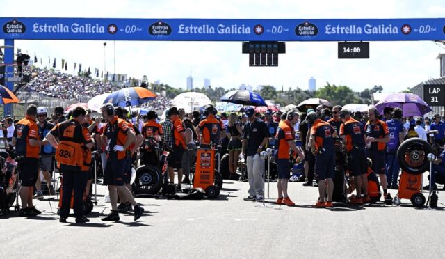 Pit crews in orange uniforms organize at the starting grid under an Estrella Galicia banner, with spectators behind them and race equipment scattered across the track.