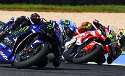 Group of professional motorcycle racers leaning into a tight turn on a race track, blue Yamaha in foreground with Monster Energy branding.