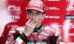 MotoGP rider in a red racing suit touches his lips in a thoughtful pose during a press event, with sponsor logos on a red backdrop behind him.