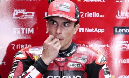 MotoGP rider in a red racing suit touches his lips in a thoughtful pose during a press event, with sponsor logos on a red backdrop behind him.