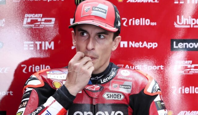MotoGP rider in a red racing suit touches his lips in a thoughtful pose during a press event, with sponsor logos on a red backdrop behind him.