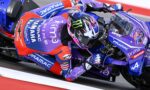 MotoGP rider in purple Pramac livery leans into a turn on a blue race bike, helmet with pink and blue graphics.