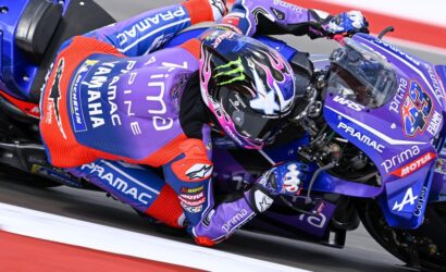 MotoGP rider in purple Pramac livery leans into a turn on a blue race bike, helmet with pink and blue graphics.