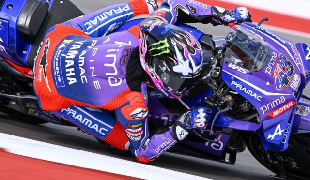 MotoGP rider in purple Pramac livery leans into a turn on a blue race bike, helmet with pink and blue graphics.