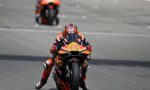 MotoGP rider in orange and blue racing suit leaning into a turn on a high-speed track, front-wheel barely off the ground, bike #12 with Red Bull livery ahead of blurred bike in background