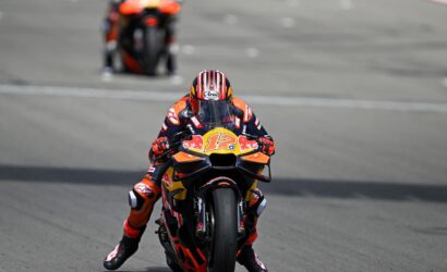 MotoGP rider in orange and blue racing suit leaning into a turn on a high-speed track, front-wheel barely off the ground, bike #12 with Red Bull livery ahead of blurred bike in background