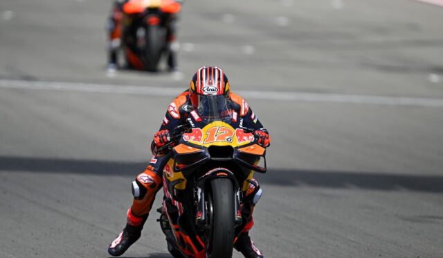 MotoGP rider in orange and blue racing suit leaning into a turn on a high-speed track, front-wheel barely off the ground, bike #12 with Red Bull livery ahead of blurred bike in background