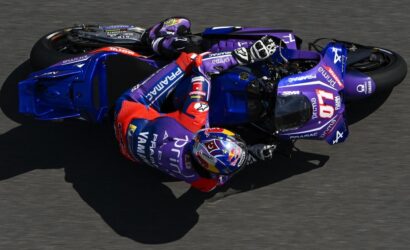 Motorcycle racer in purple racing suit leaning into a sharp turn on a blue Yamaha race bike on a track, helmeted and in full gear.