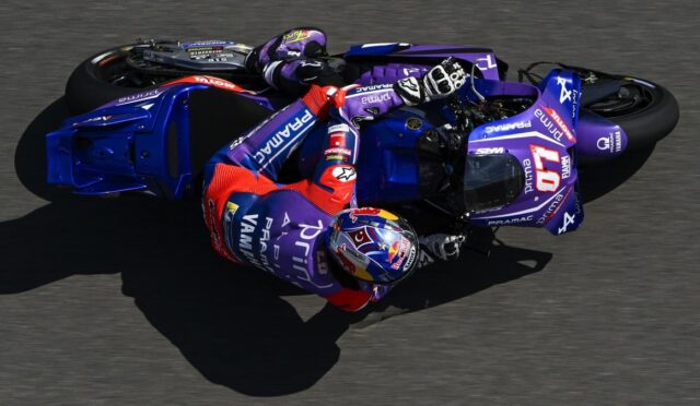 Motorcycle racer in purple racing suit leaning into a sharp turn on a blue Yamaha race bike on a track, helmeted and in full gear.