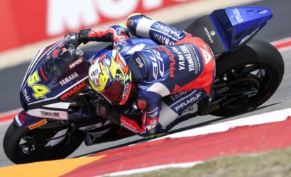 MotoGP rider in a blue-red Pramac Yamaha suit leaning into a turn on a blue Yamaha motorcycle; bright yellow helmet visible.