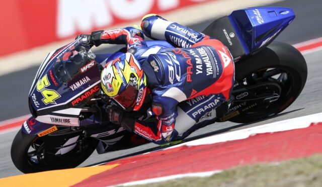 MotoGP rider in a blue-red Pramac Yamaha suit leaning into a turn on a blue Yamaha motorcycle; bright yellow helmet visible.