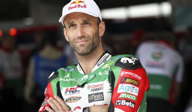 Male motorcycle racer in a green sponsorship suit and a white Red Bull cap, arms crossed and looking toward the camera in a pit area.