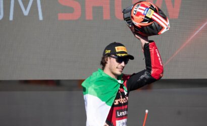 Racing rider raises helmet in celebration on stage, wearing a black Pirelli cap and Italian flag cape behind a large display screen.