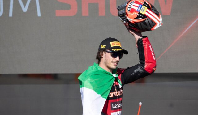Racing rider raises helmet in celebration on stage, wearing a black Pirelli cap and Italian flag cape behind a large display screen.