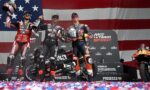 Three MotoGP riders on the winners' podium celebrating with drink bottles, backed by a large American flag banner and sponsor boards. The center rider holds a trophy or helmet, with two others raising their bottles in celebration.