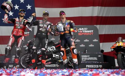 Three MotoGP riders on the winners' podium celebrating with drink bottles, backed by a large American flag banner and sponsor boards. The center rider holds a trophy or helmet, with two others raising their bottles in celebration.